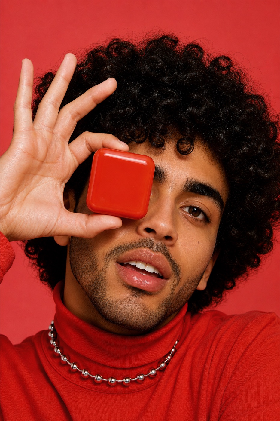 Young man against a red background holding a red solid perfume compact in front of his eye.
